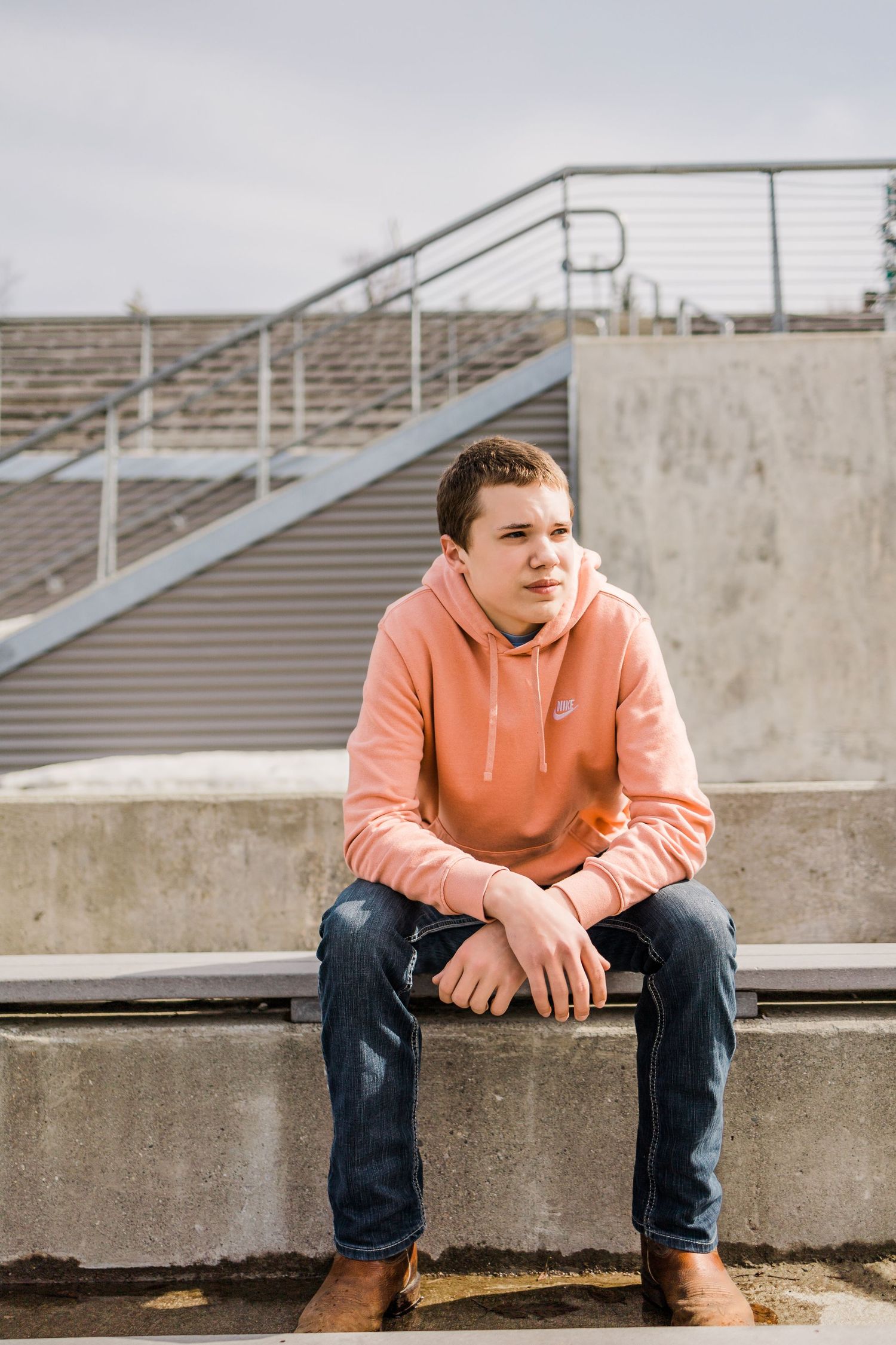 Teenage boy in a peach Nike hoodie sits on concrete bleachers at an outdoor stadium.