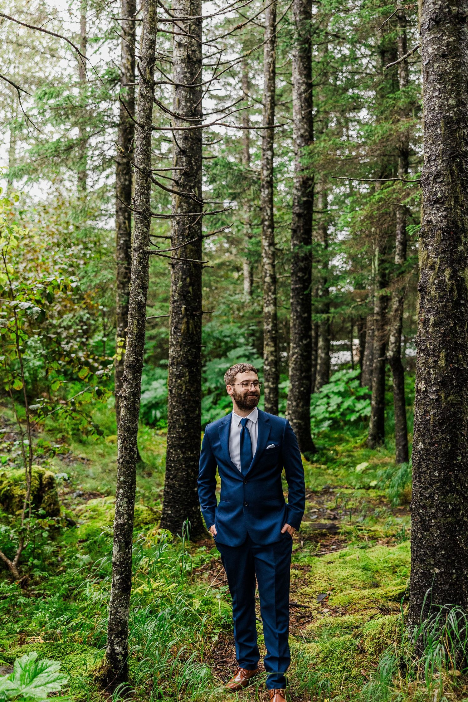 Groom in navy suit stands among tall evergreen trees in mossy forest setting.