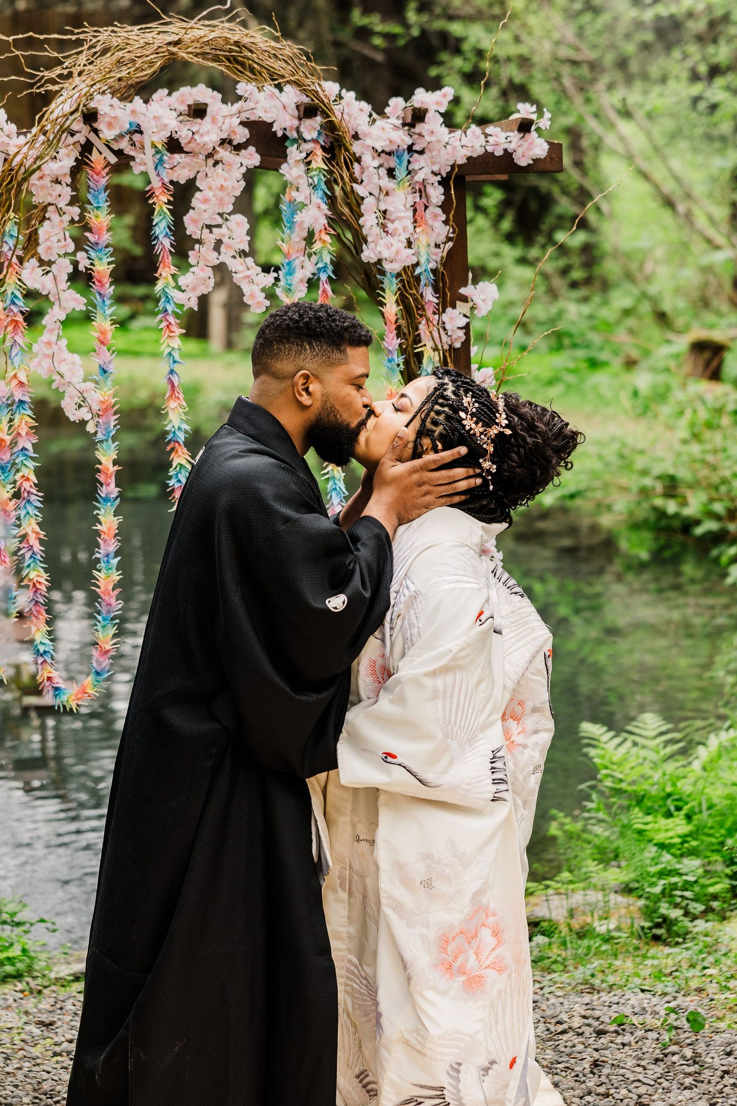 Couple shares romantic moment under floral archway by forest pond.
