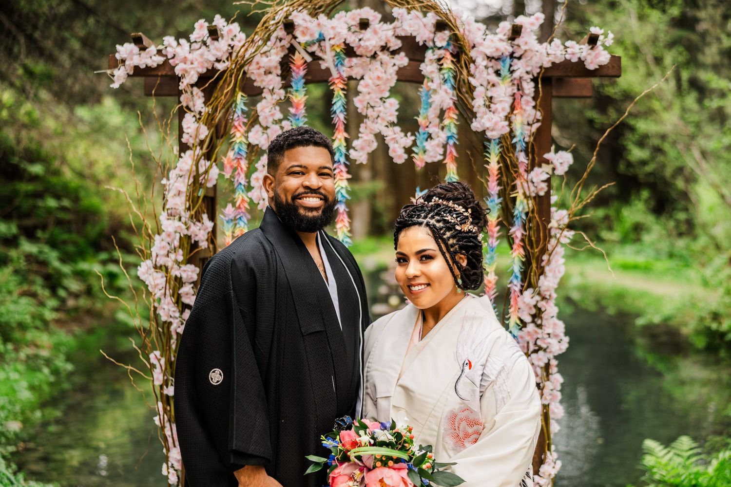 Wedding couple standing under a floral arch decorated with pink cherry blossoms in a forest setting.
