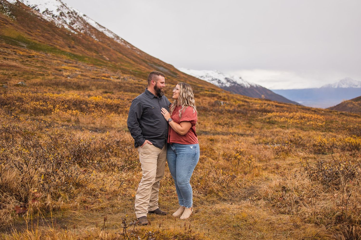 Couple standing together in golden autumn tundra with snow-dusted mountains in the distance.