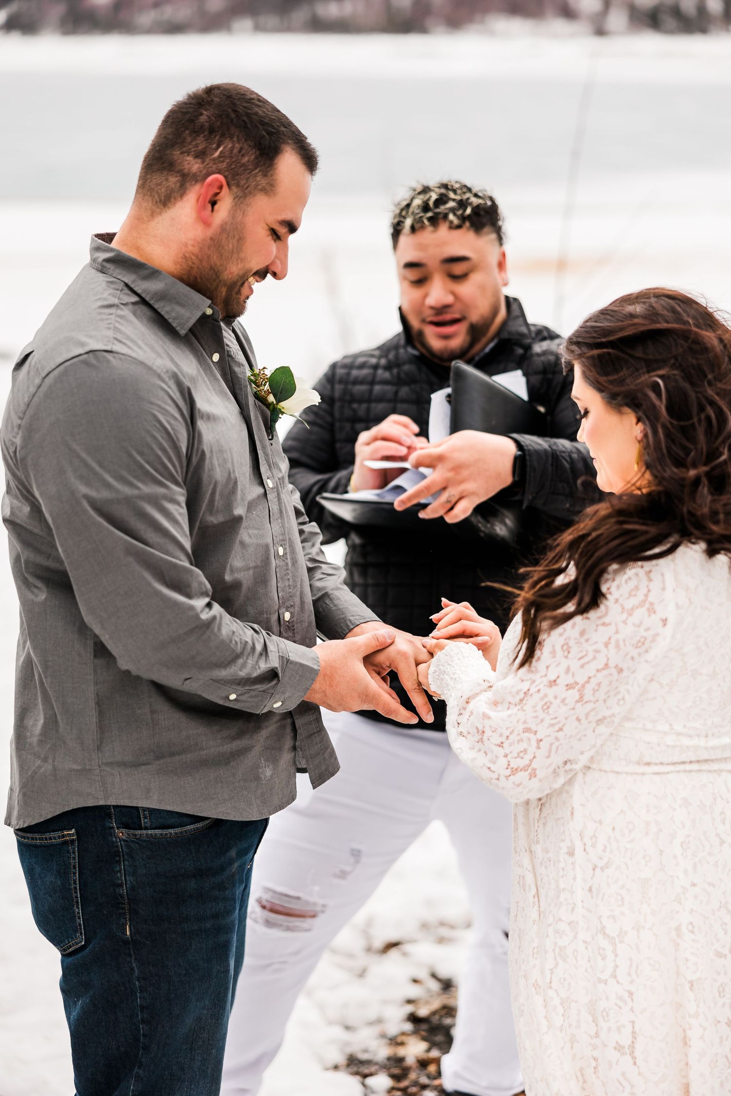 Groom smiles as bride places wedding ring on his finger during intimate snowy outdoor ceremony.