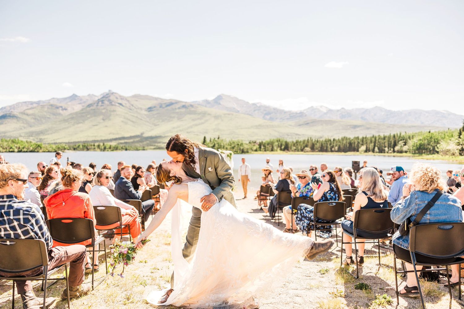 Wedding guests seated at outdoor ceremony venue with mountains and lake in background.