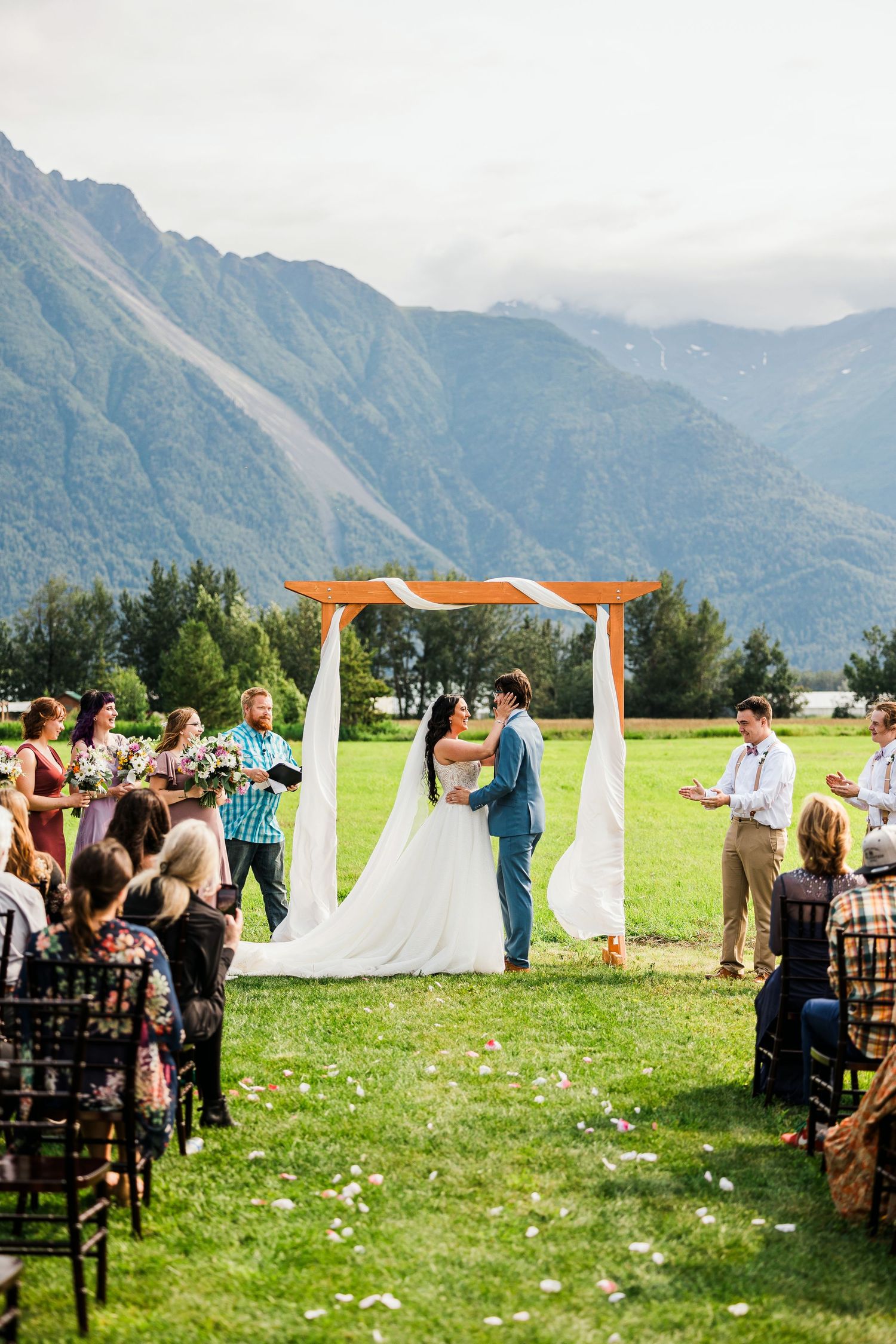 Wedding ceremony in progress under wooden arch with dramatic mountain landscape in background.