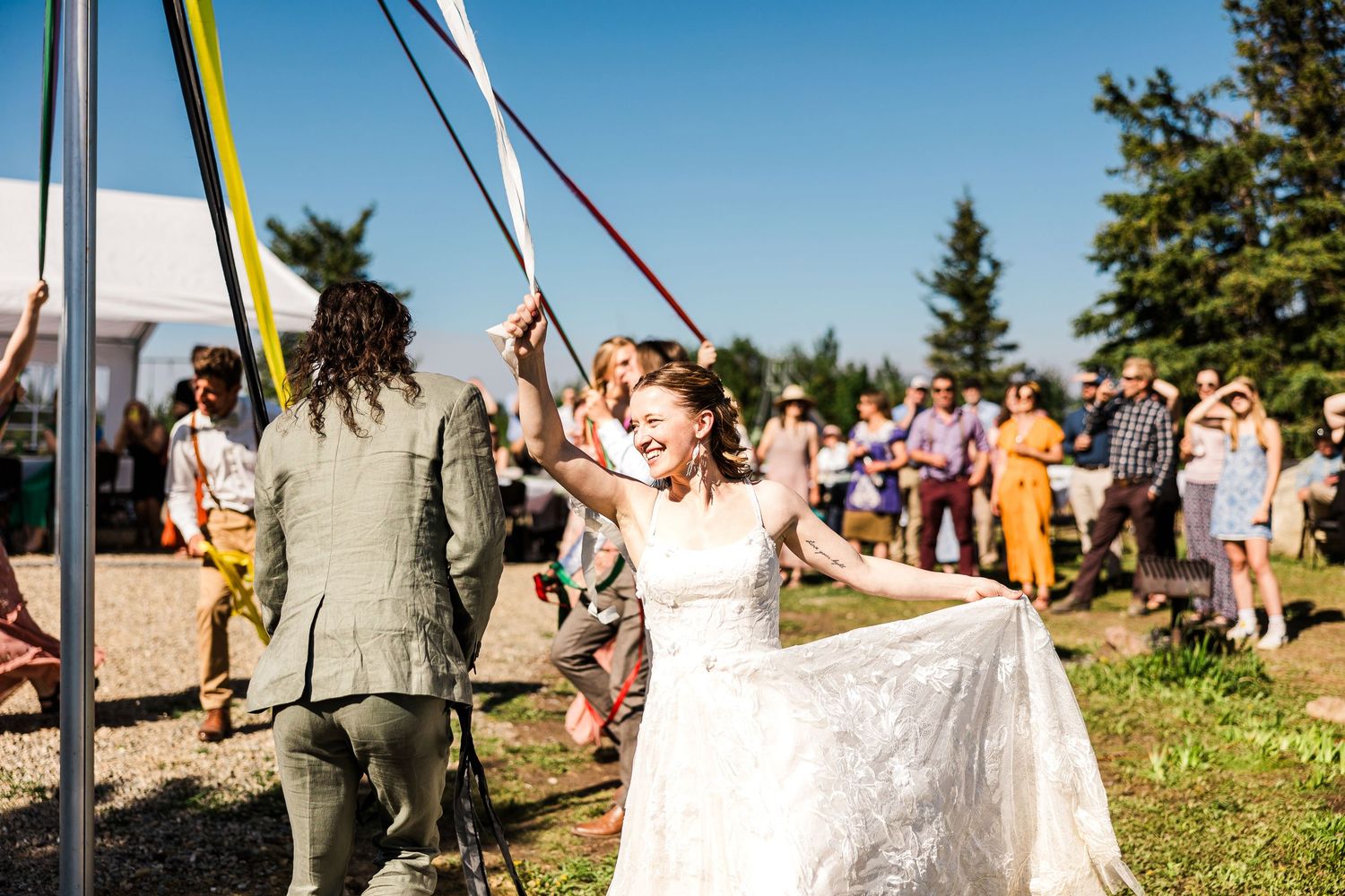 Outdoor wedding celebration with guests gathered around a maypole under blue skies.