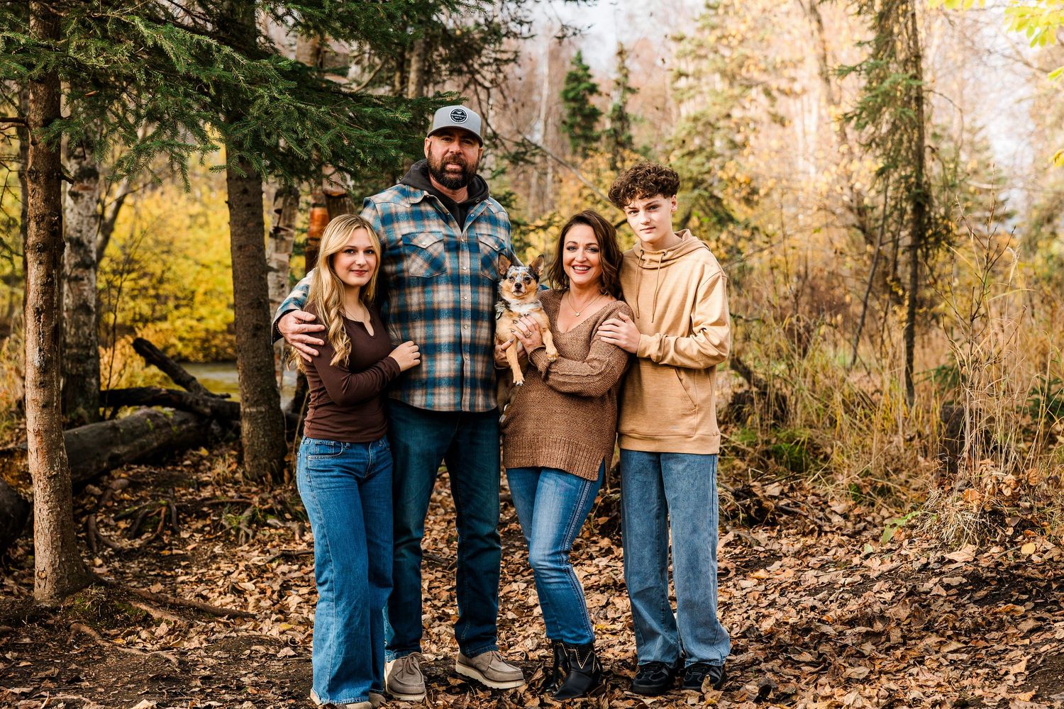A group of people wearing casual fall attire pose together in a wooded area with autumn foliage.