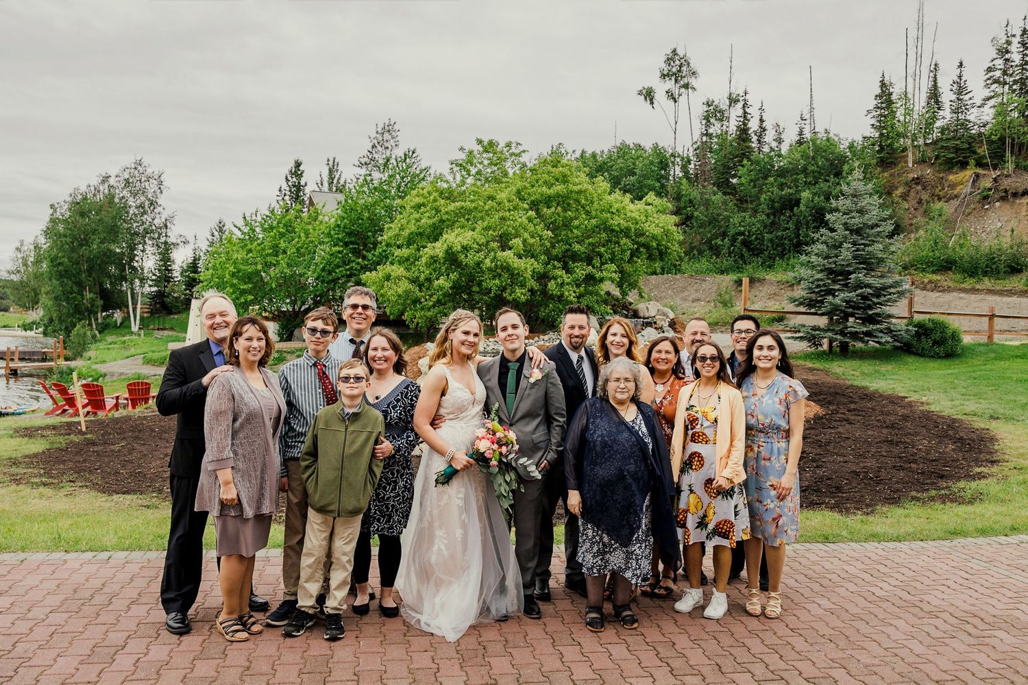 Wedding party poses outdoors on brick patio surrounded by lush green trees at elegant garden venue.
