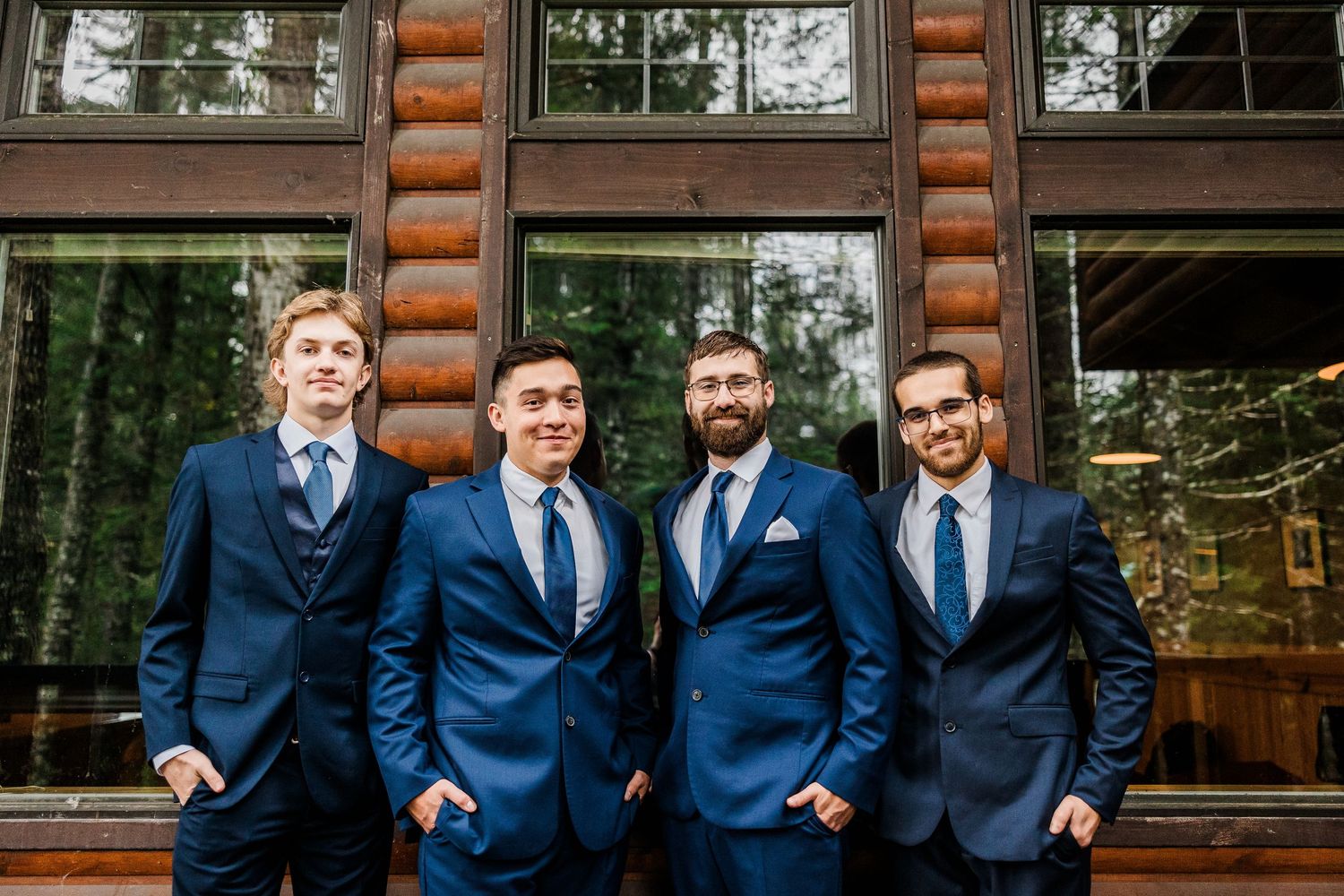 Groomsmen in matching navy blue suits pose in front of a rustic log cabin.