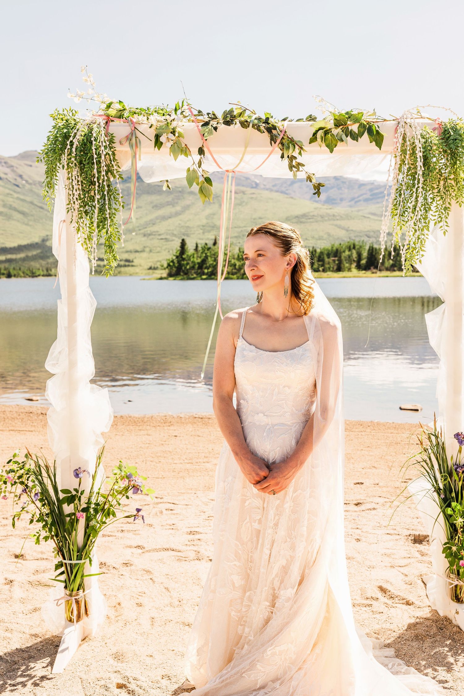 Bride in lace gown poses under floral arch with lake and mountain scenery.
