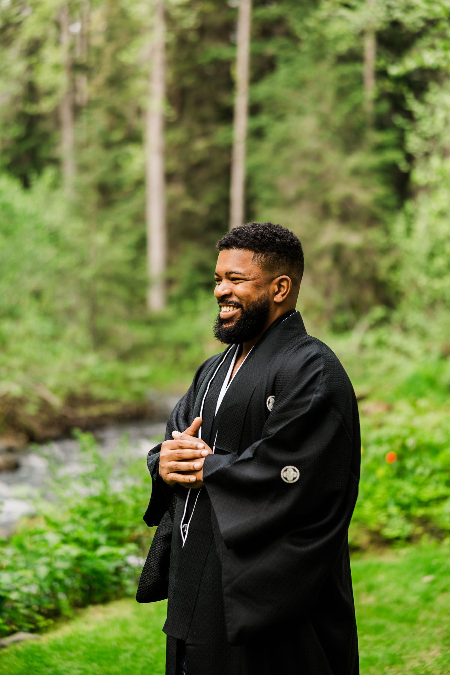 Person in traditional black kimono smiles joyfully by forest stream.
