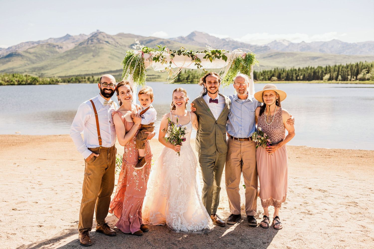 Wedding party poses on beach with mountain lake backdrop and decorated arch.