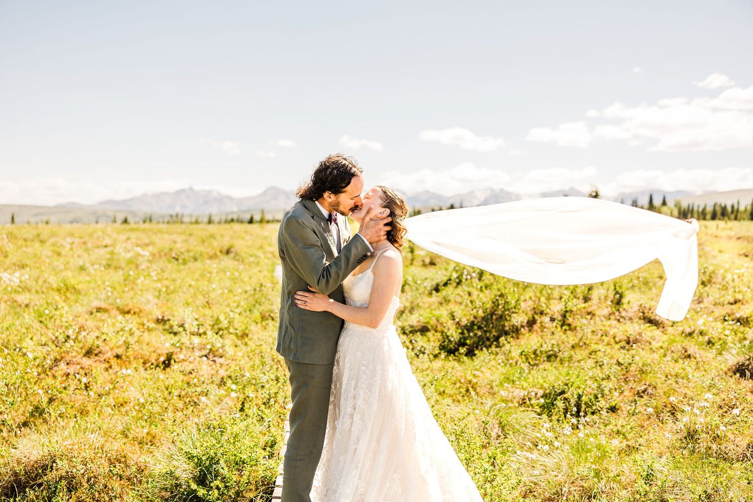 Bride and groom share a romantic kiss in a golden meadow with mountains and flowing veil.