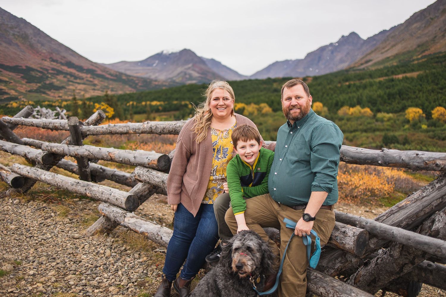 Couple with young son and black dog posing at rustic log fence in mountain valley.