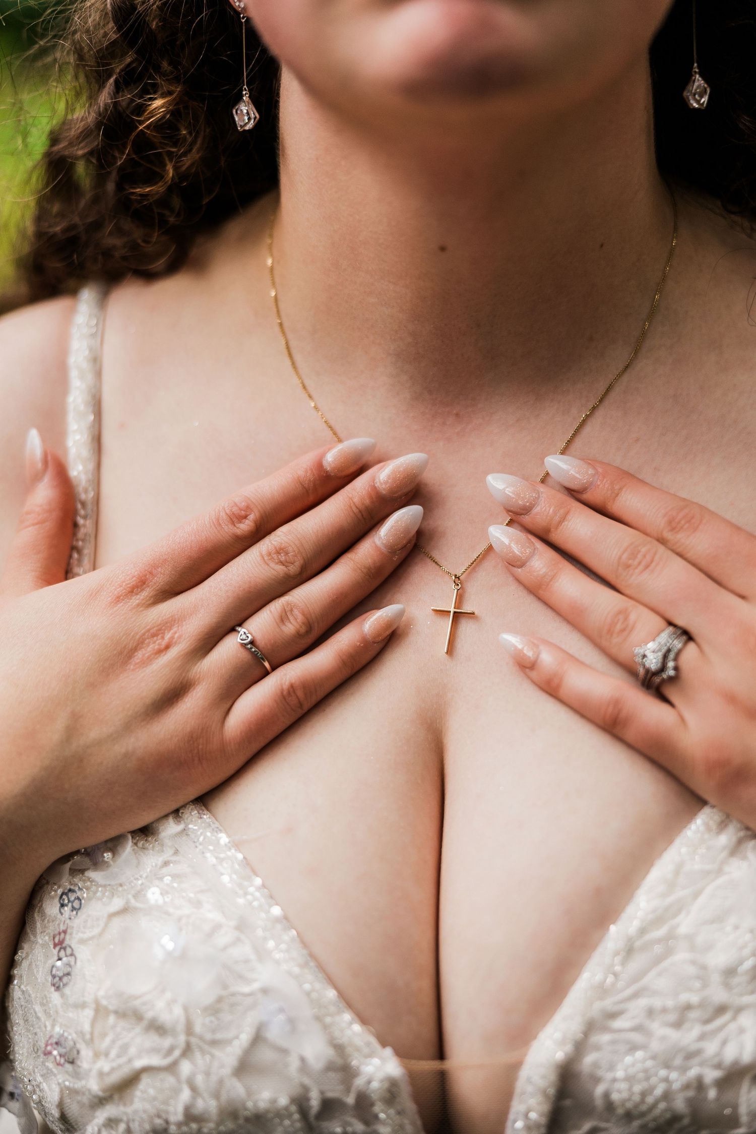 Close up of bride's jewelry and lace neckline with hands showing rings.