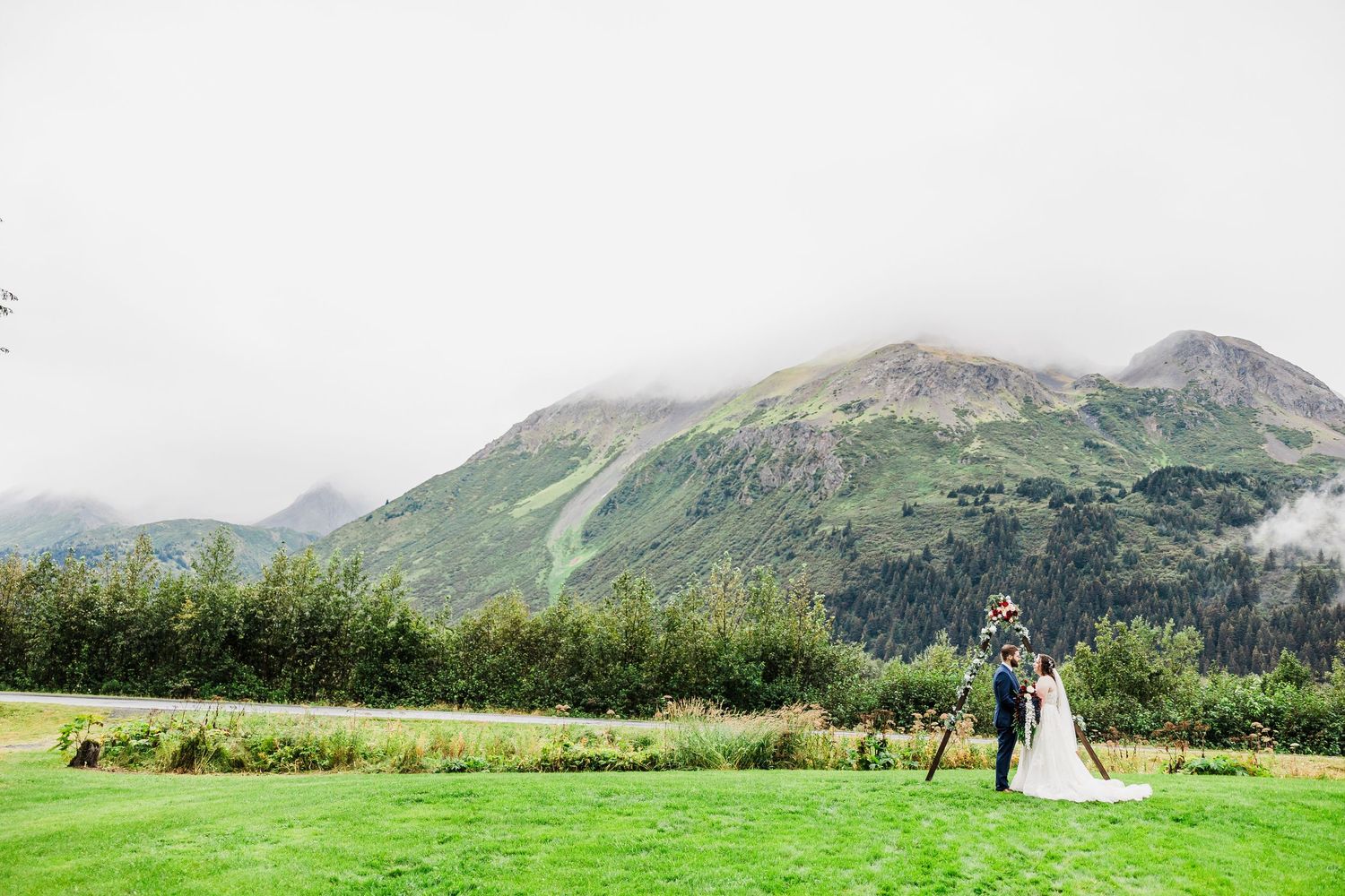 Bride and groom standing at altar arch on lush green lawn with misty mountains.