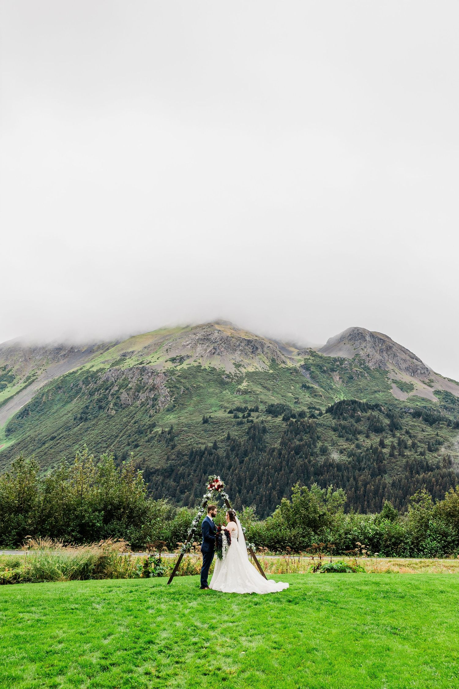 Wedding couple stands isolated on green meadow with misty mountain backdrop.
