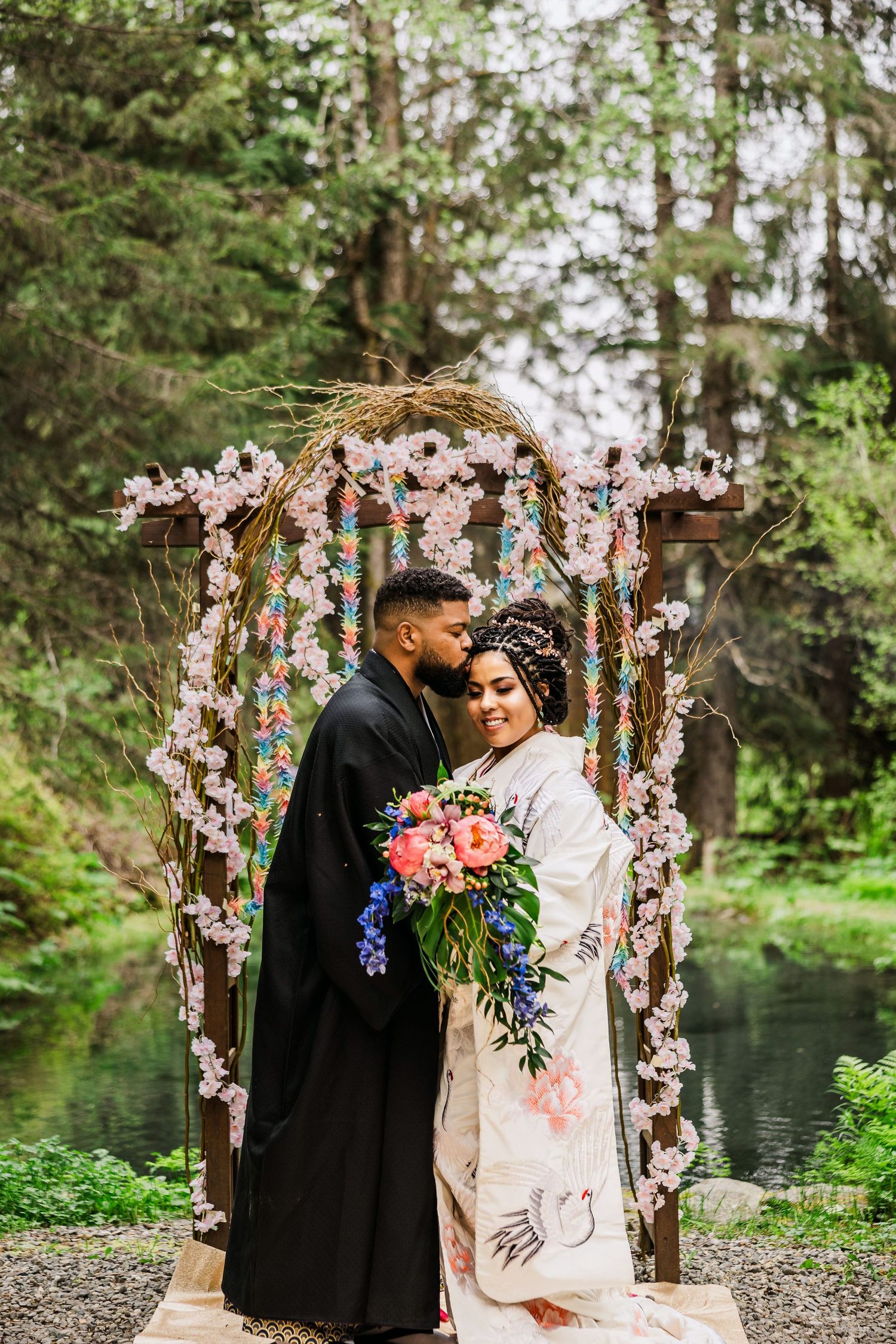 Couple in traditional Japanese wedding attire embrace before floral arch in lush forest setting.