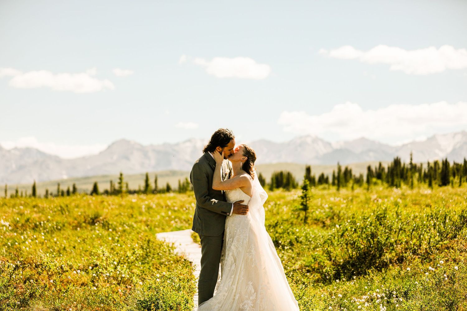 Bride and groom share a romantic kiss in a vibrant yellow wildflower field with mountain views.