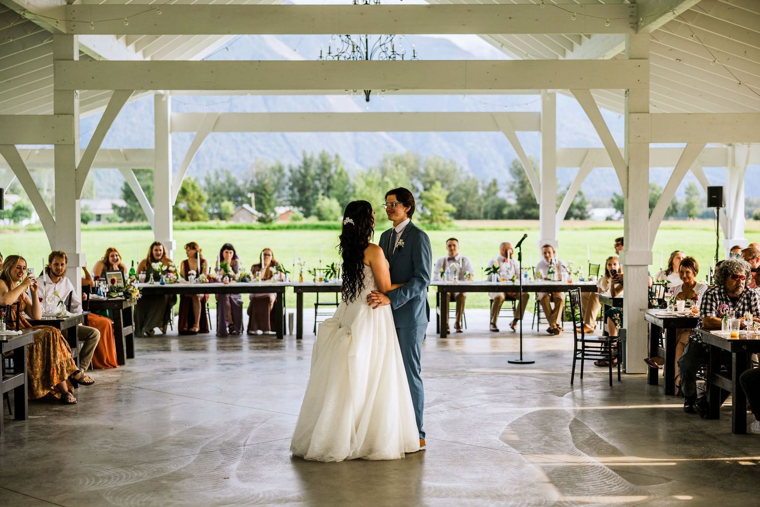 Couple shares their first dance at wedding reception under white pavilion while guests watch from tables.