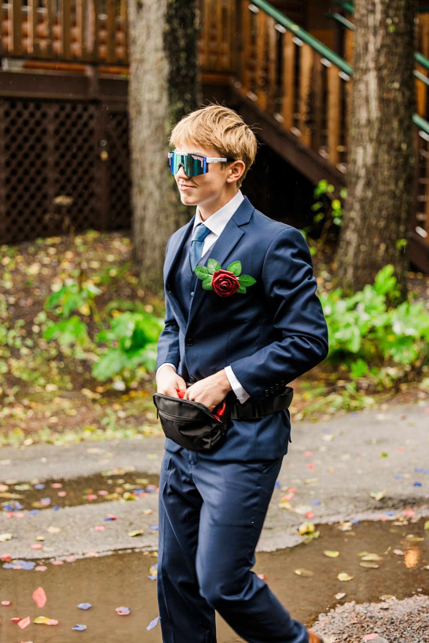 A person in a navy suit and blue sunglasses with a red boutonniere walks outdoors.