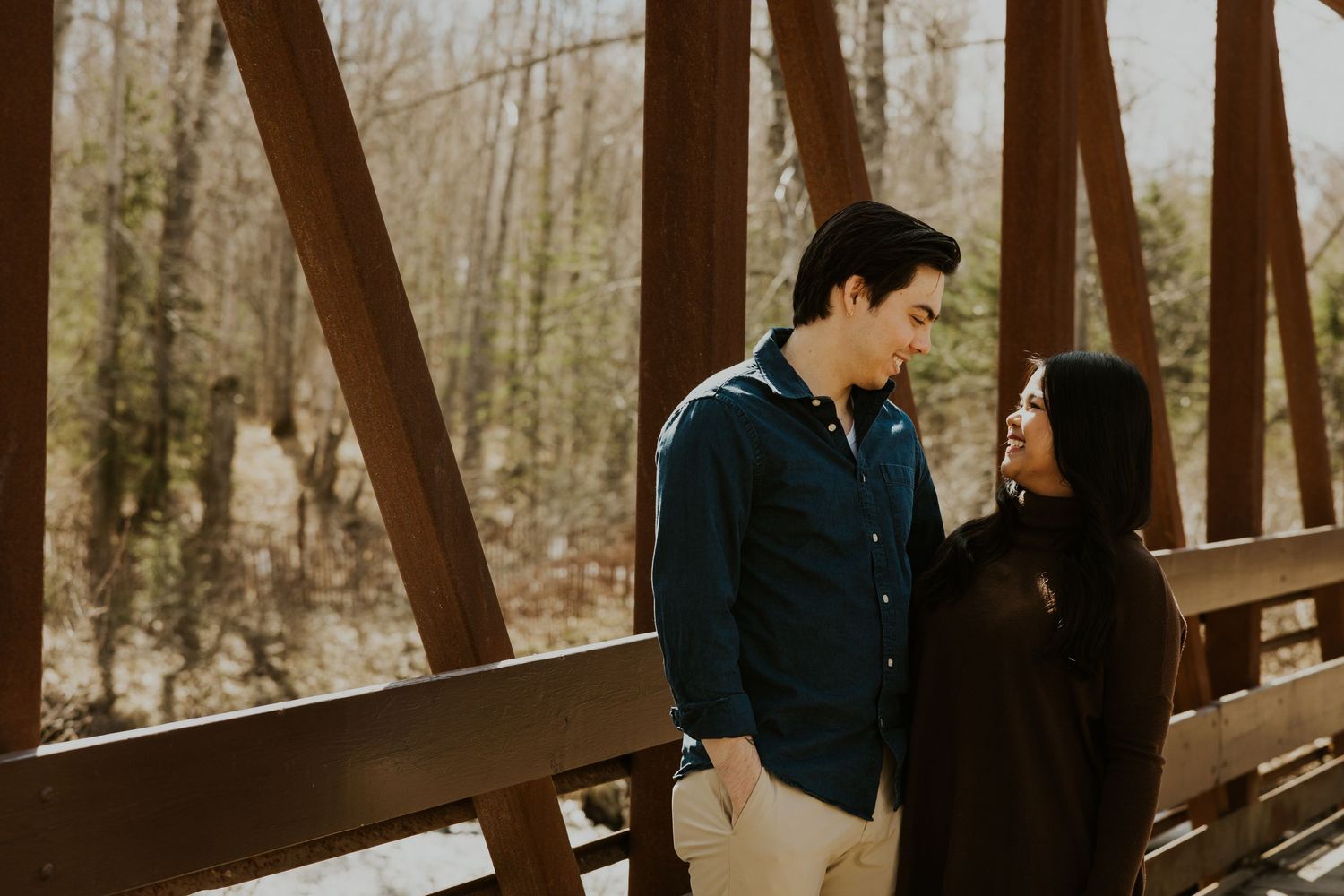 Young couple sharing a smile on a wooden bridge surrounded by bare winter trees.