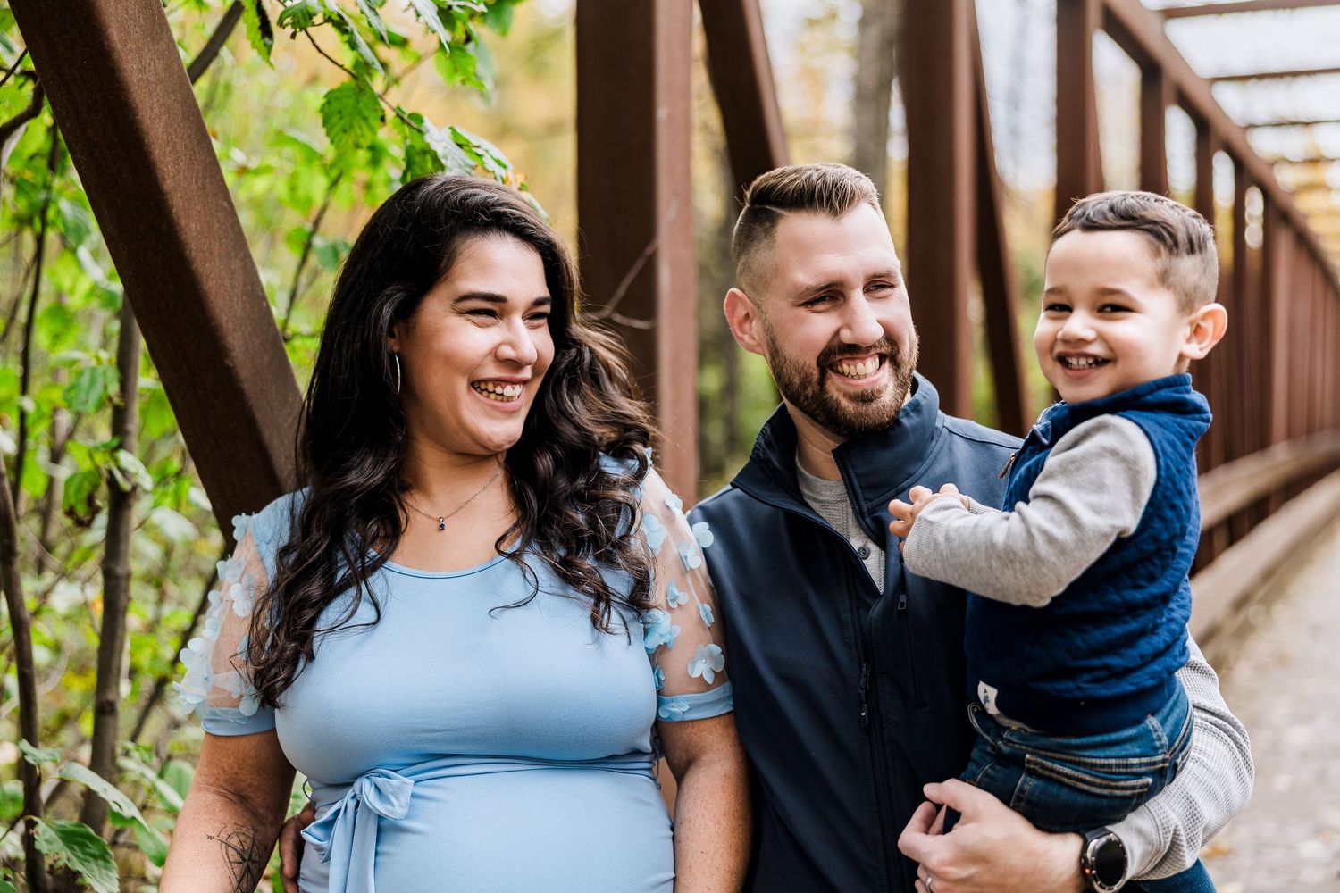 Happy family portrait with parents and toddler son smiling on a bridge in fall foliage.