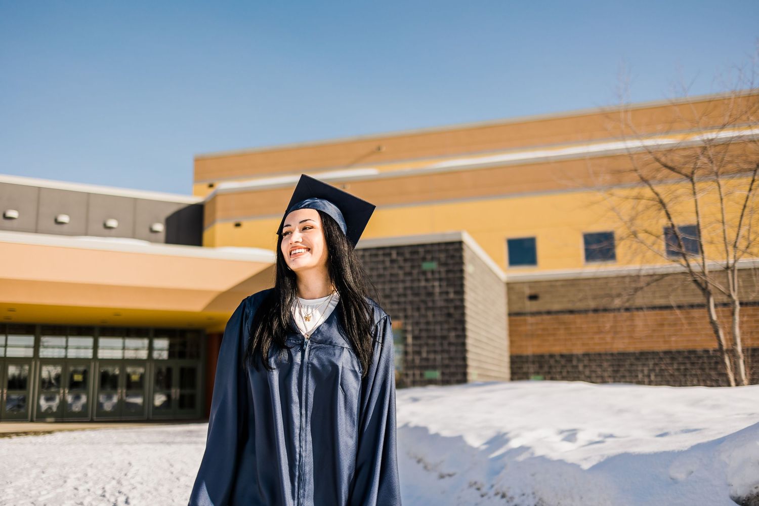 Smiling graduate in cap and gown stands outside a school building in a snowy winter setting.