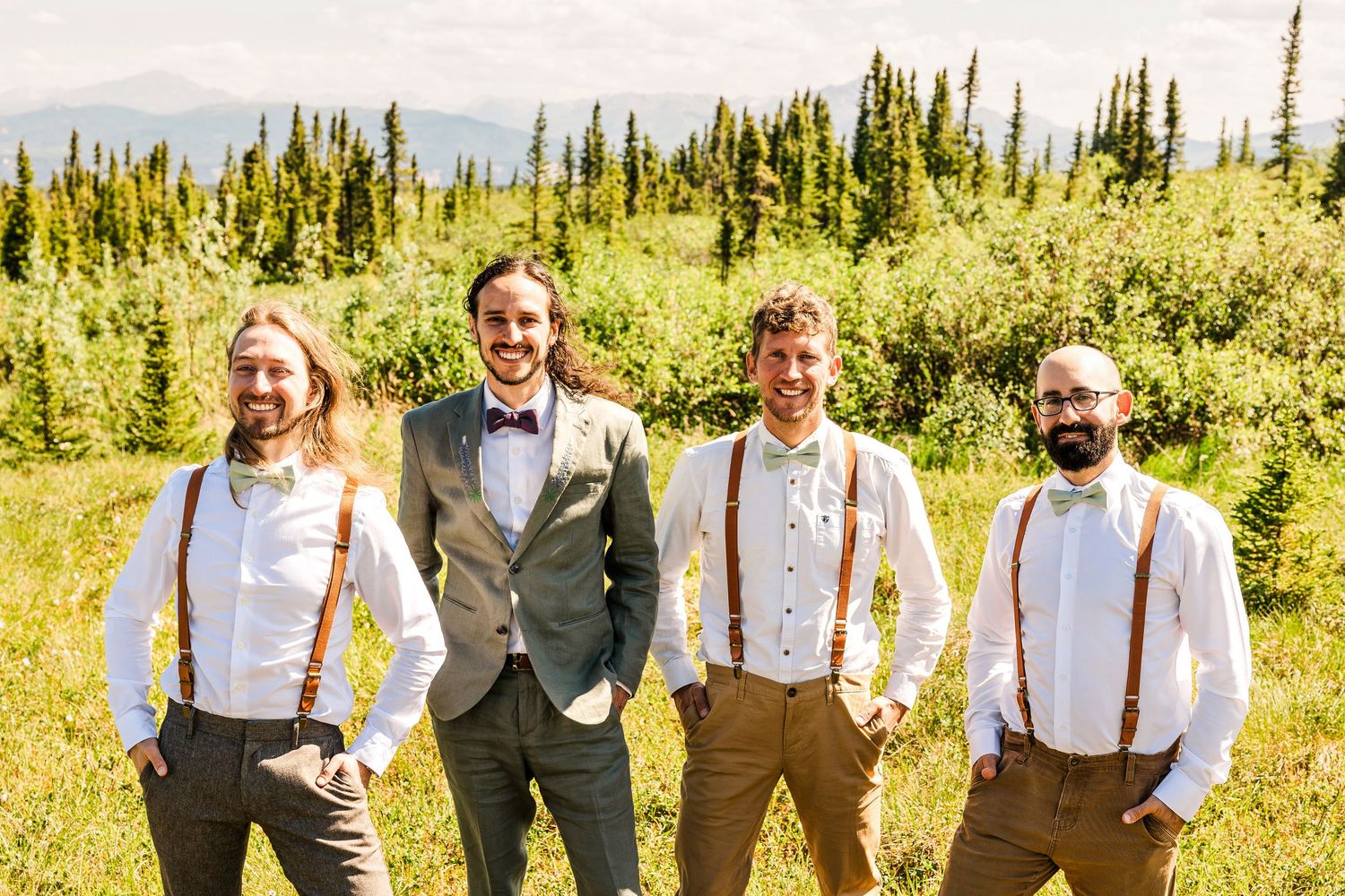 Groomsmen in suspenders and bowties pose together in sunny outdoor setting.