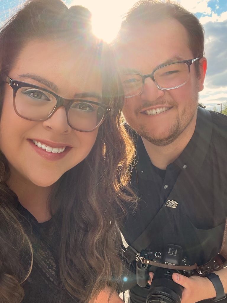 Smiling couple wearing glasses takes a sunny selfie together while holding a professional camera.