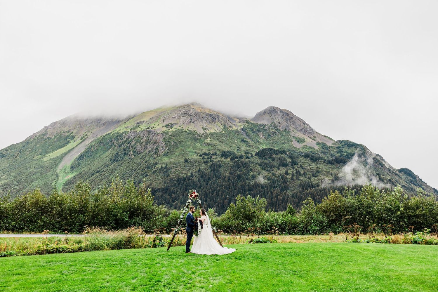 Bride and groom stand under floral arch on green lawn with misty mountain backdrop at outdoor wedding.