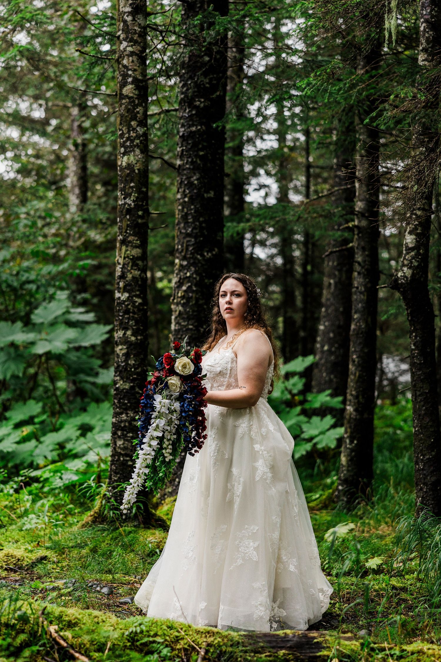 Bride in lace gown poses with bouquet in enchanted forest setting.