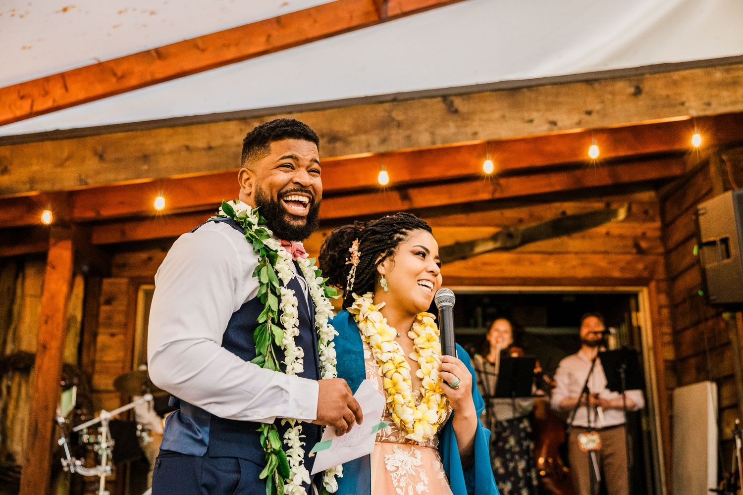 A couple wearing traditional Hawaiian leis share a joyful moment during their wedding reception in a rustic venue.