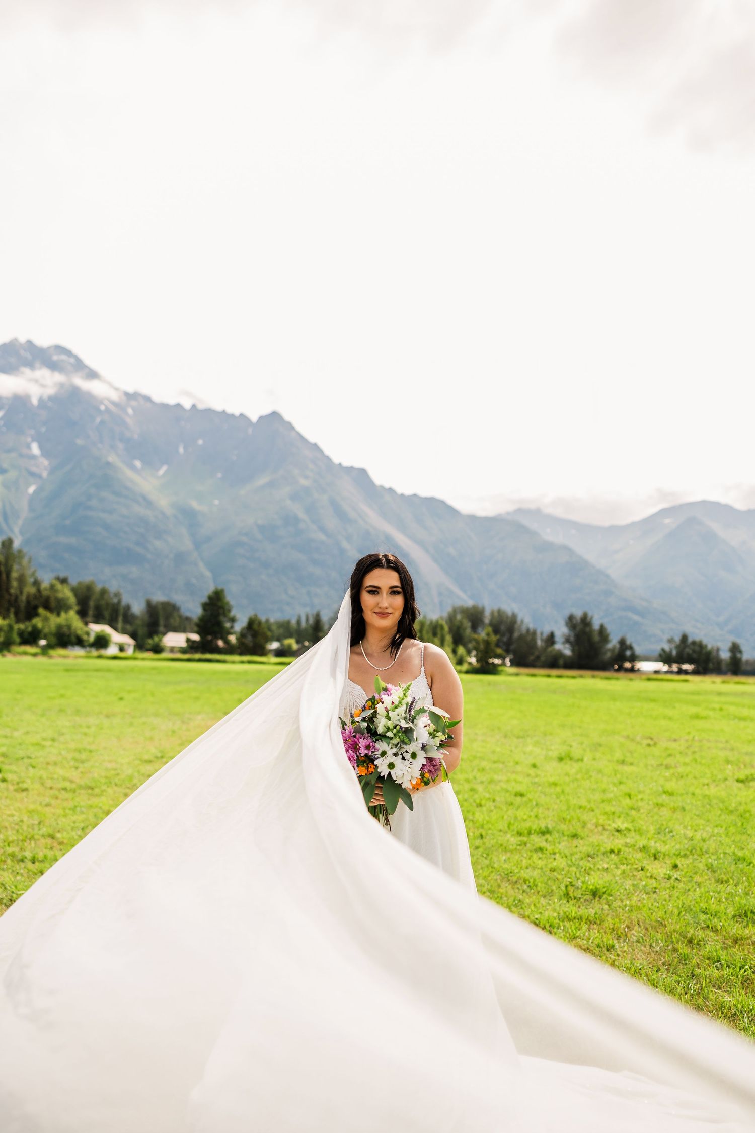 Bride in flowing white gown and veil holds colorful bouquet in green meadow with mountain backdrop.