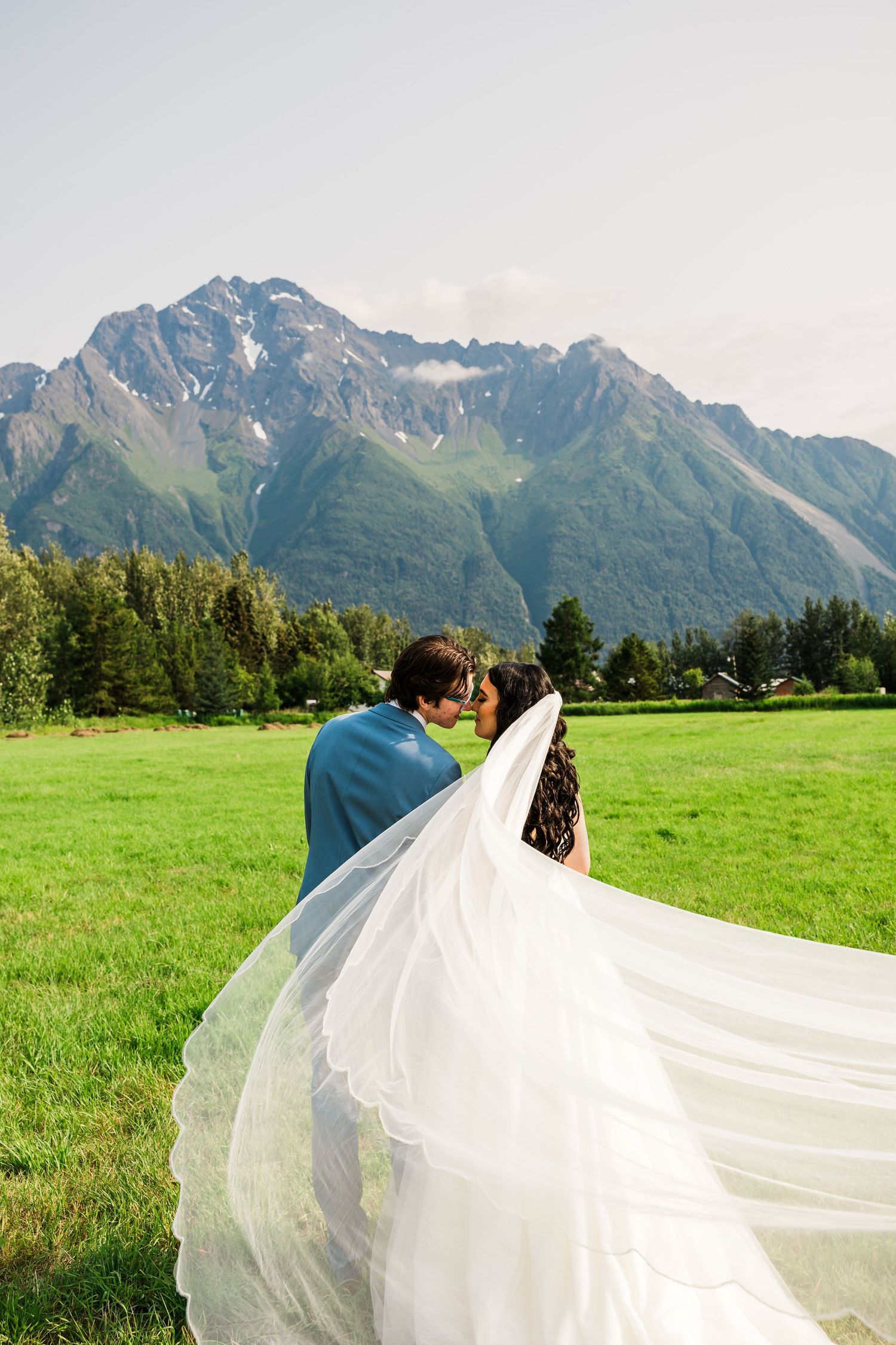 Long flowing veil catches breeze as couple embraces in mountain meadow setting.