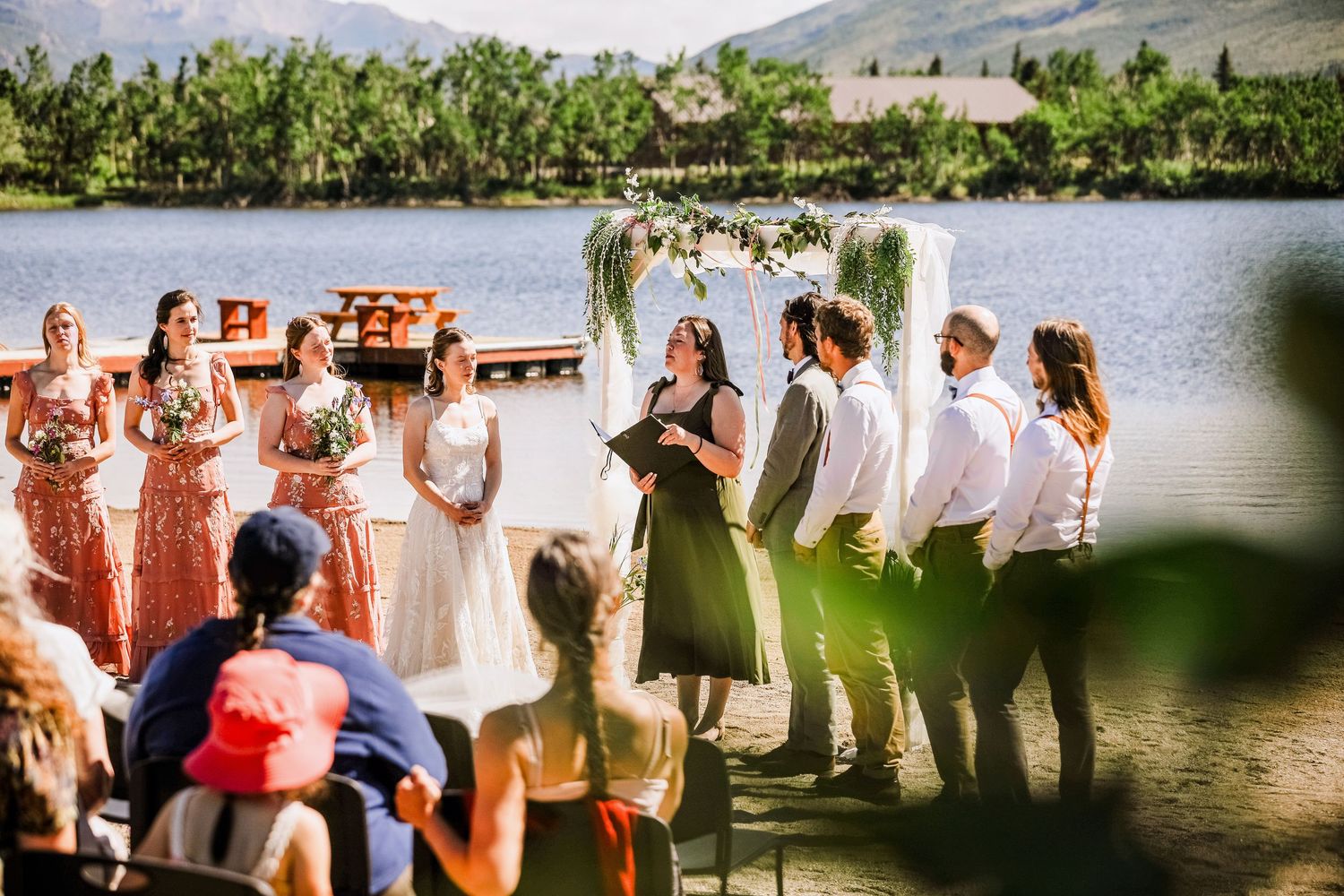 Lakeside dock wedding ceremony with wedding party and guests gathered under floral arch.