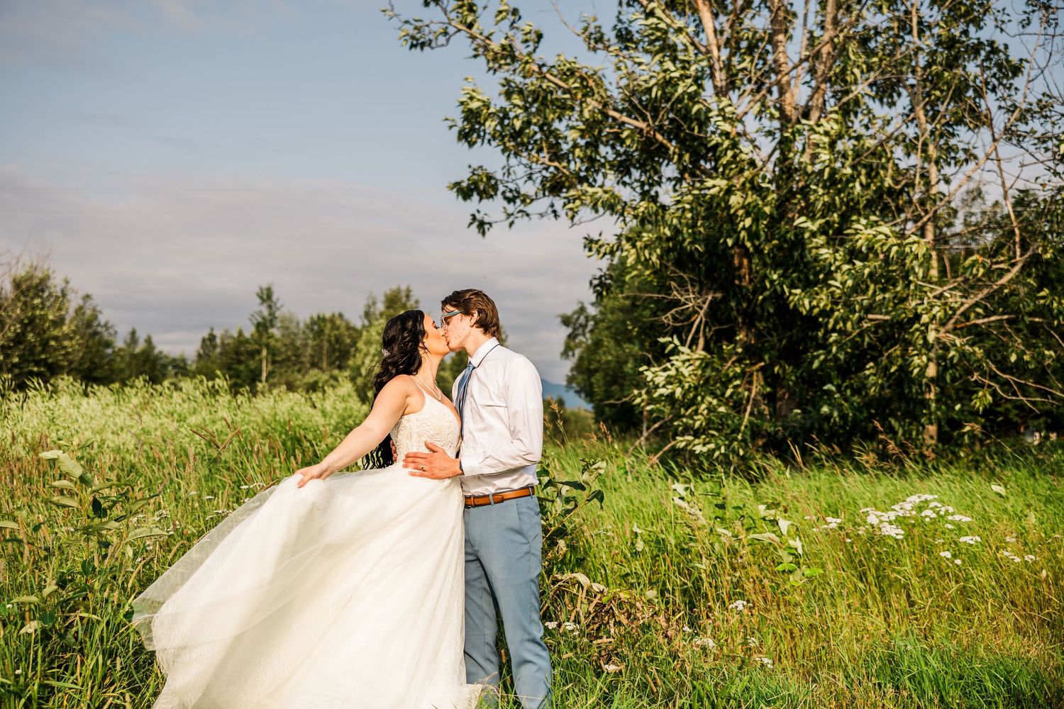 Newlyweds share a romantic moment in a sunlit meadow with mountains in the background.