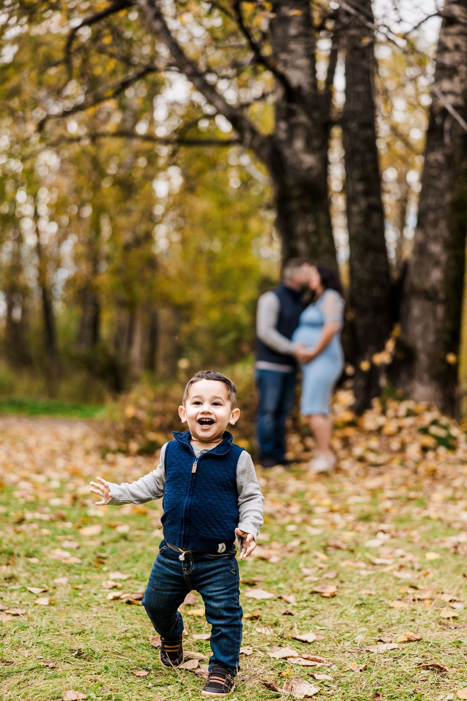 Excited toddler runs toward camera while expecting parents embrace near autumn trees behind him.