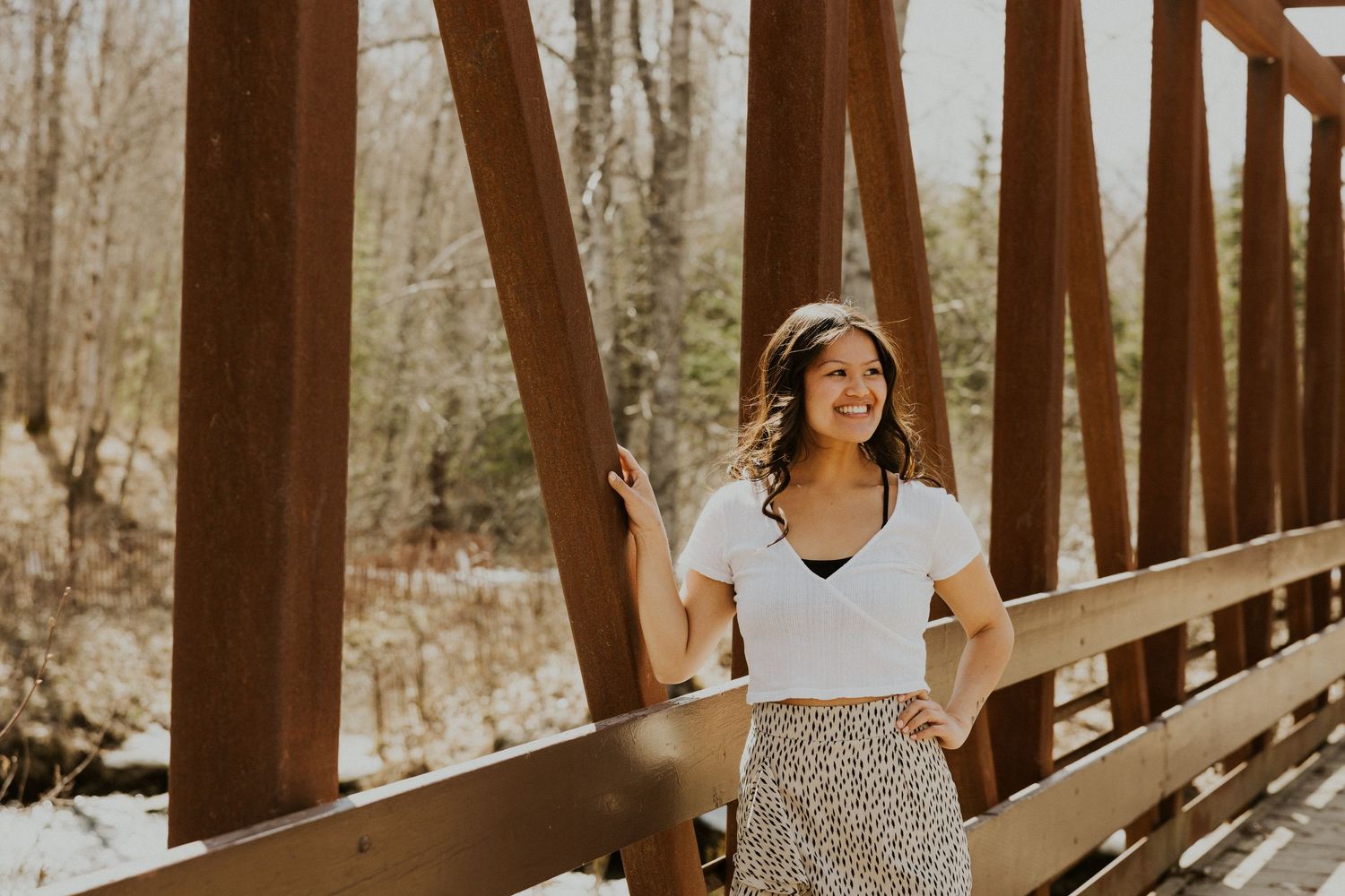 Smiling young woman in a white top and floral skirt poses on a rustic wooden bridge in winter.