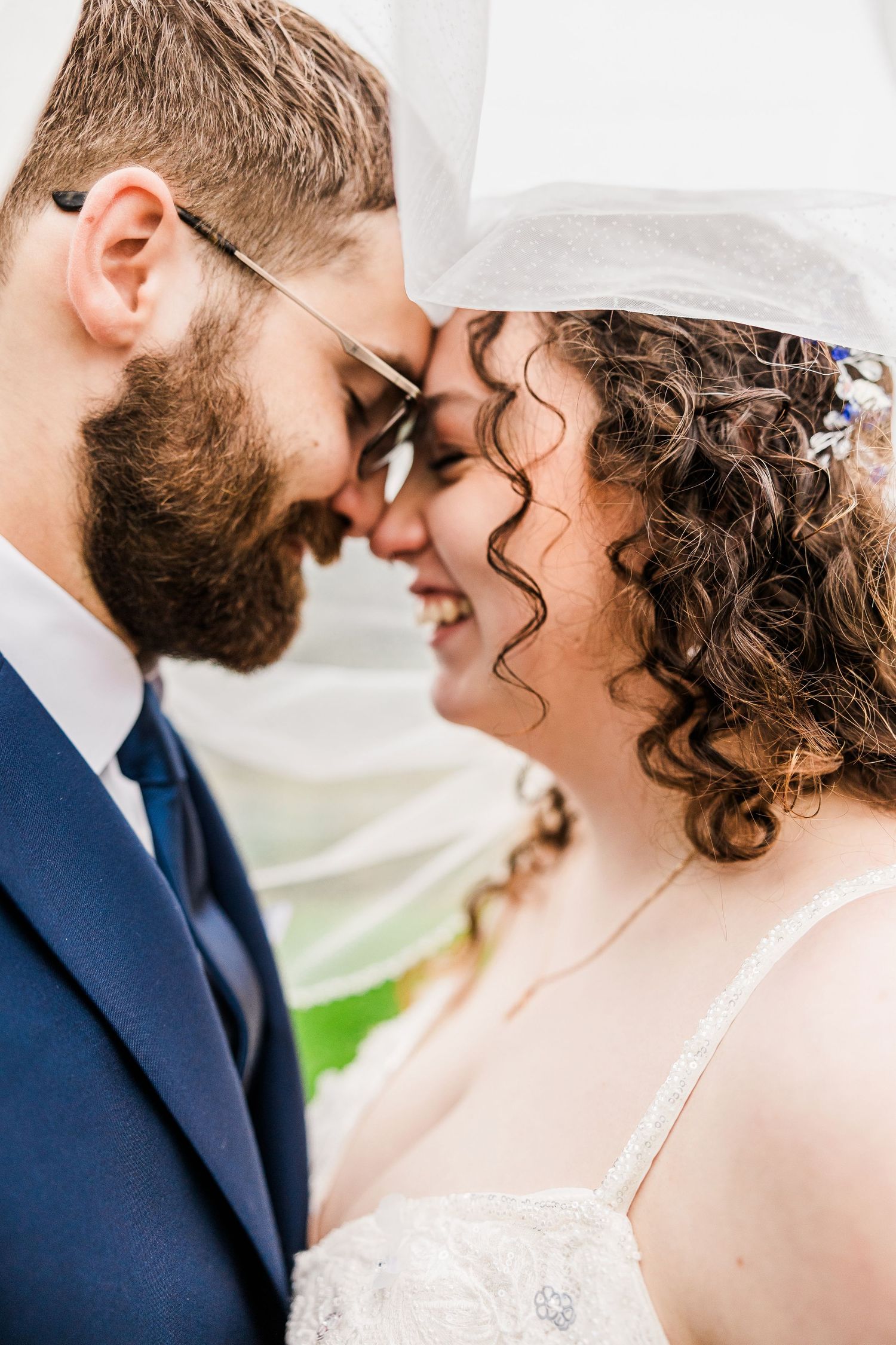Close-up of smiling bride and bearded groom touching foreheads tenderly under wedding veil.
