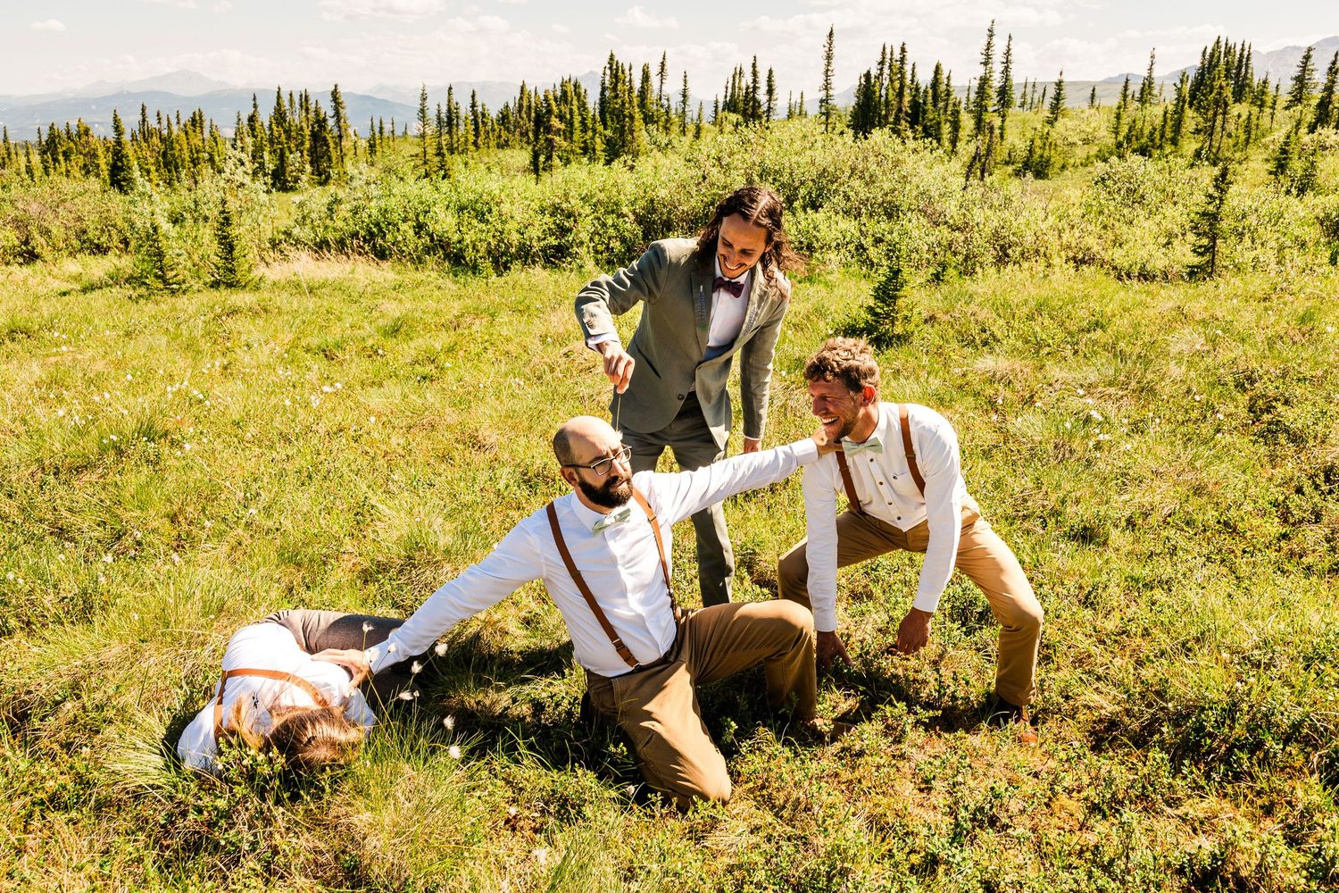 Wedding party lounges casually in a scenic mountain meadow with evergreen trees in the background.