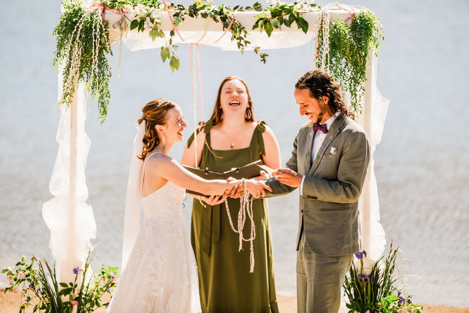 Wedding ceremony with officiant under a floral arch decorated with greenery at an outdoor lakeside venue.