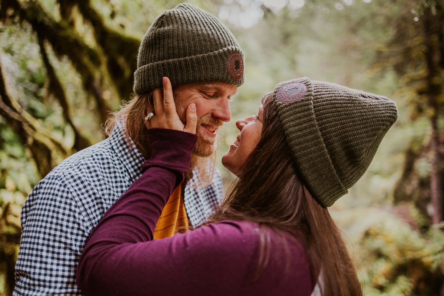 Smiling couple in matching green beanies sharing a close moment in a misty forest.
