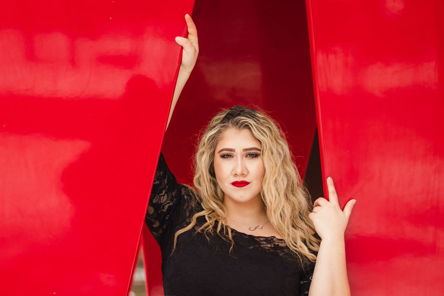 Young woman with blonde hair poses dramatically between red panels wearing a black lace top with bold red lipstick.
