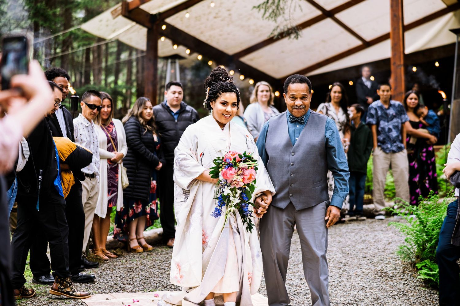 Wedding guests gather outdoors on a rustic path surrounded by greenery and trees.