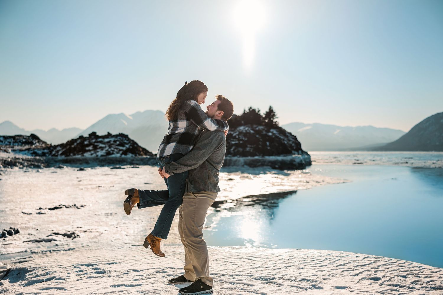 Couple embraces joyfully on a snowy beach with mountains and sparkling water in the background.