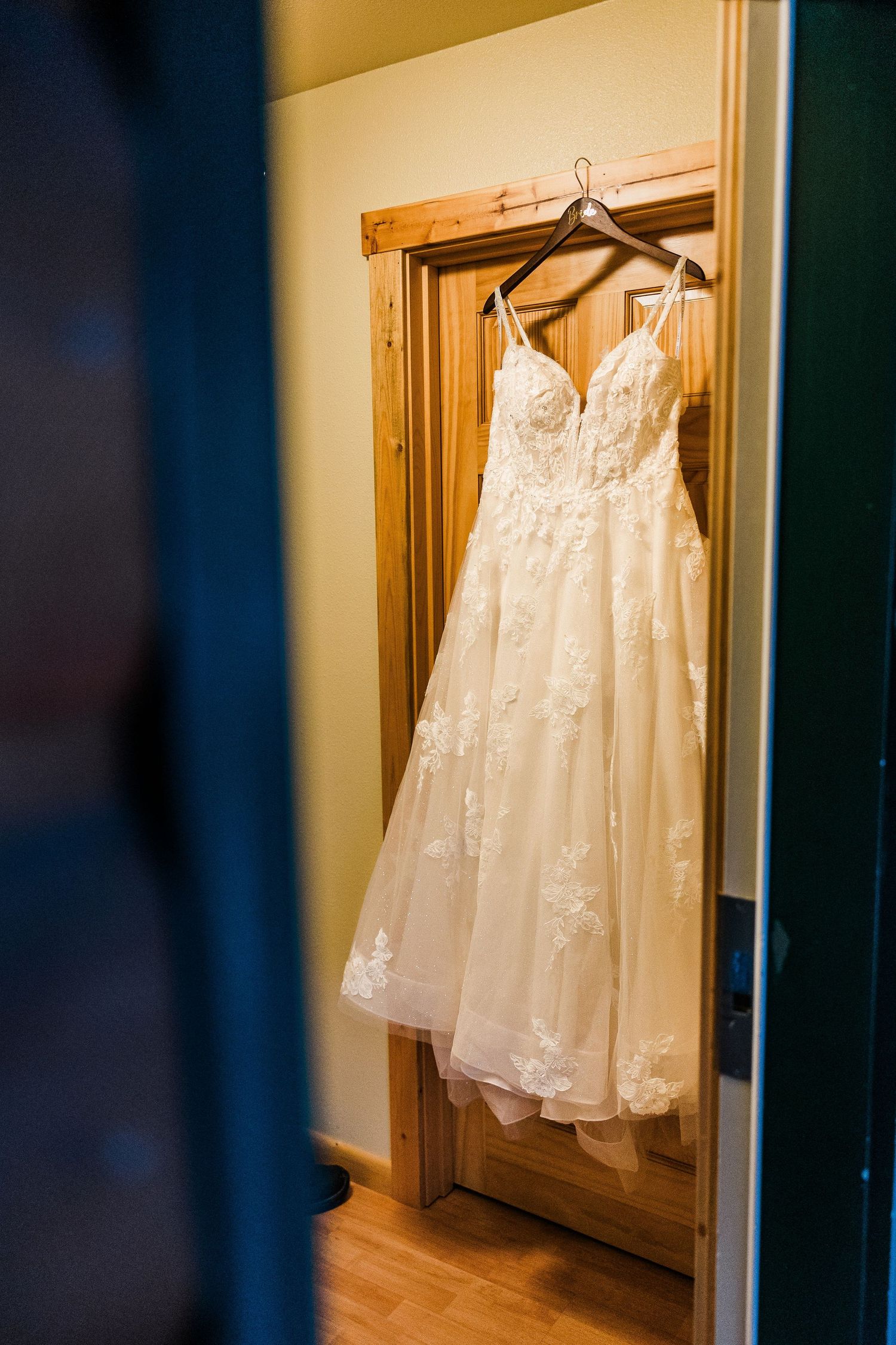 Elegant white lace wedding dress hanging on wooden door frame in natural light.