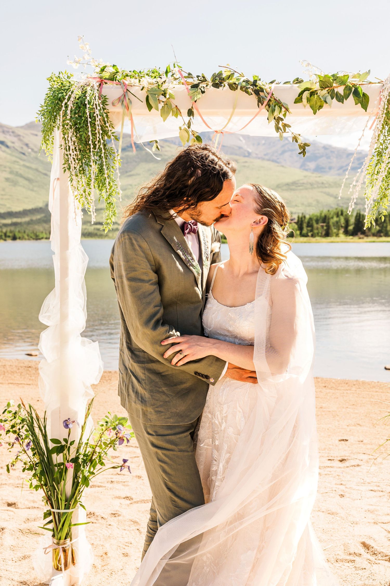 Romantic kiss under floral wedding arch with mountain lake backdrop.