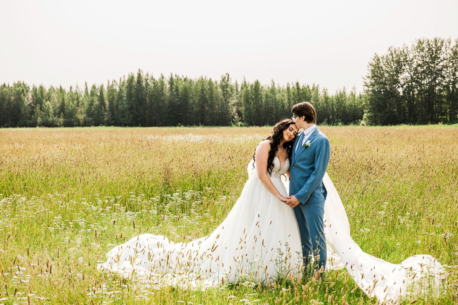 Newlyweds share intimate moment in rustic field with flowing white dress.