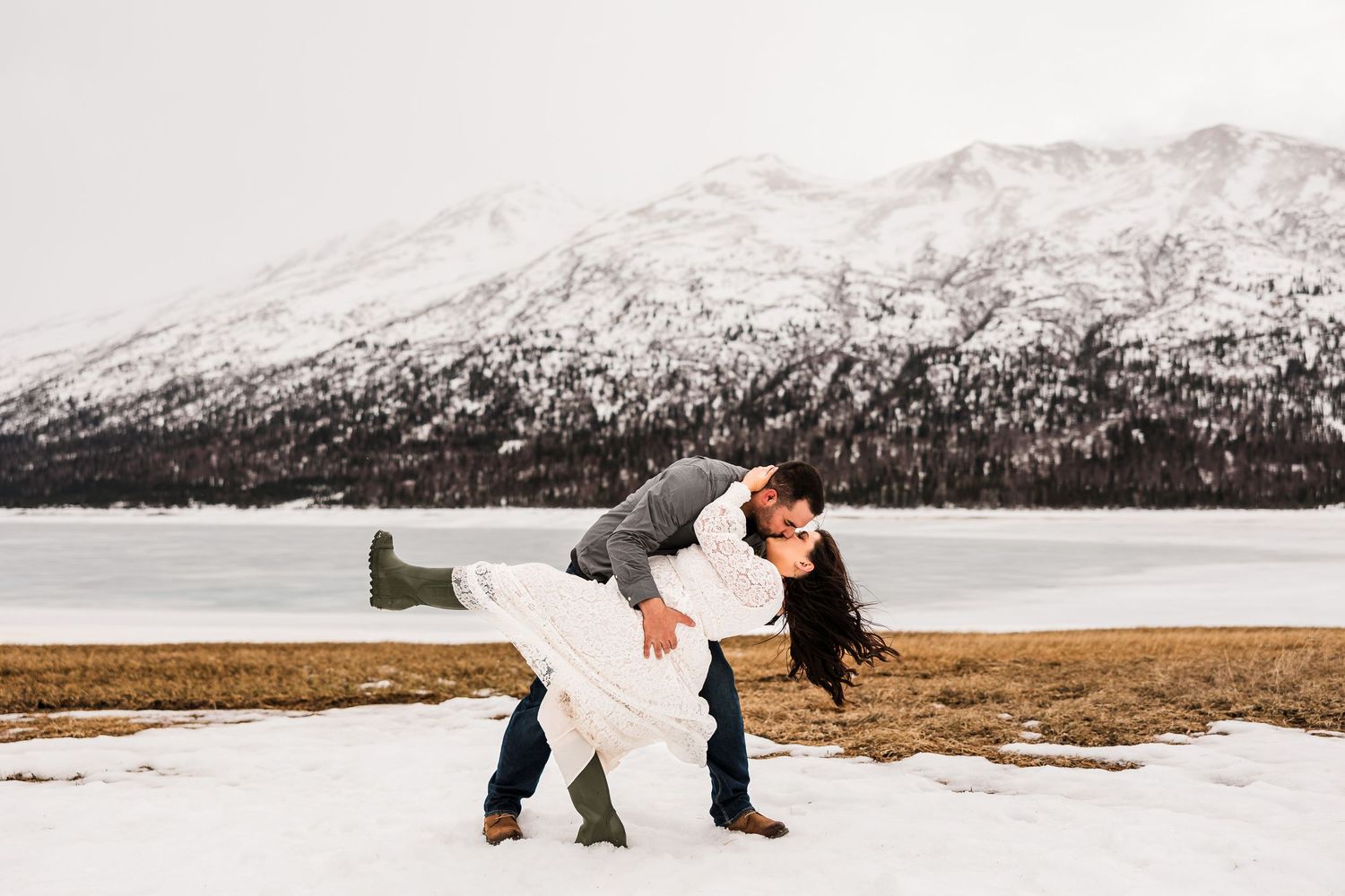 Groom dipping bride dramatically in snowy winter landscape with frozen lake and mountain.