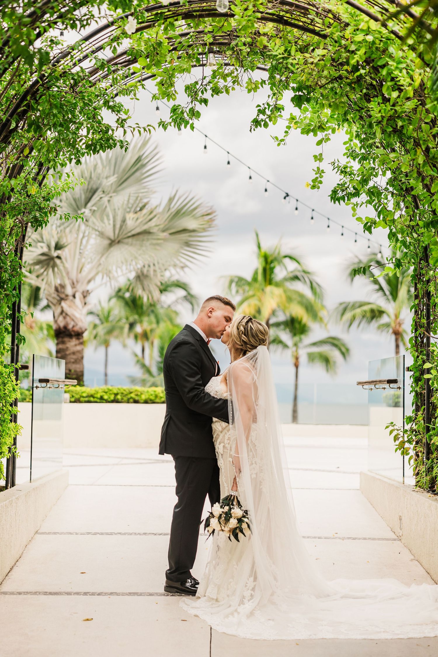 Bride and groom share a kiss under a lush green floral arch with tropical palm trees backdrop.